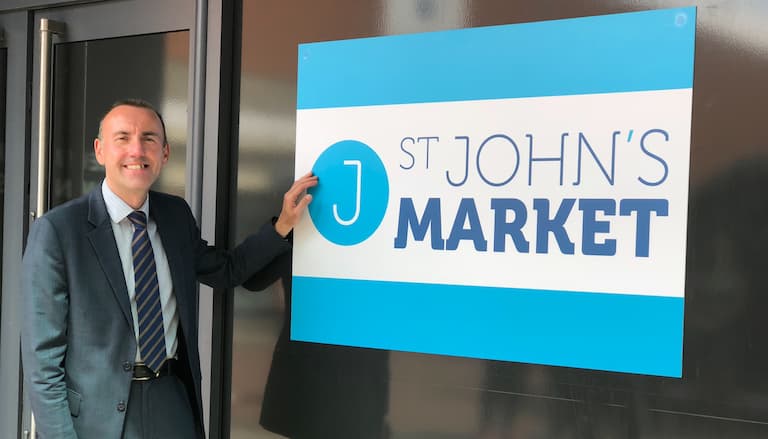 Rob Waltham smiling next to a blue and white sign for St John's Market