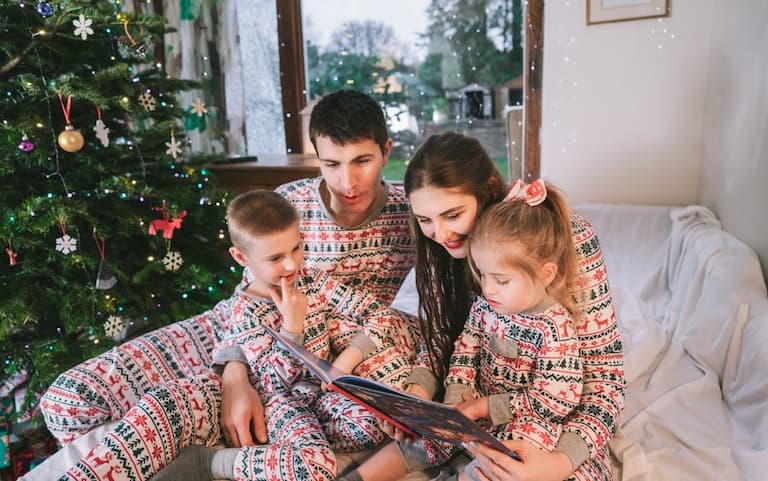 A family of four reading a book together dressed in cosy matching pyjamas next to a Christmas tree