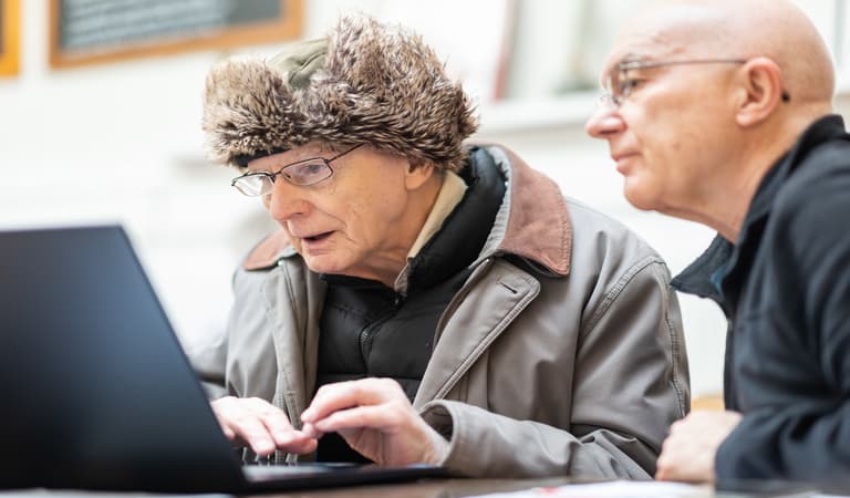 An older man in a furry hat learning how to use a laptop with another man wearing a glasses with a bald head