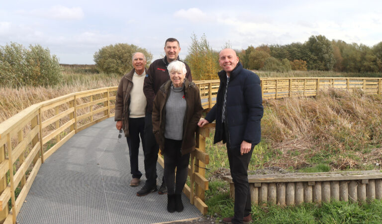 Four people standing on the boardwalk at Water's Edge surrounded by grassland