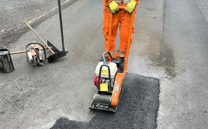 A worker in orange overalls using a machine to compact tarmac into a repaired pothole