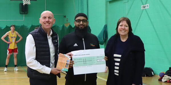 Three people holding a large cheque in a green-walled sports hall