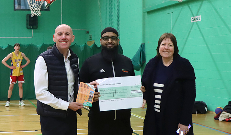 Three people holding a large cheque in a green-walled sports hall