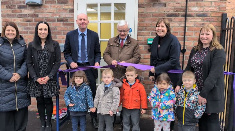 Six adults cut a purple ribbon in front of a wall. A line of small children stand in front of them.