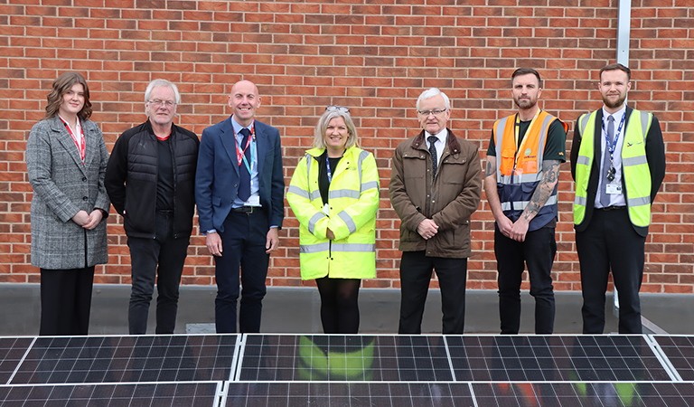 A line of people standing against a brick wall with a solar panels array in front of them