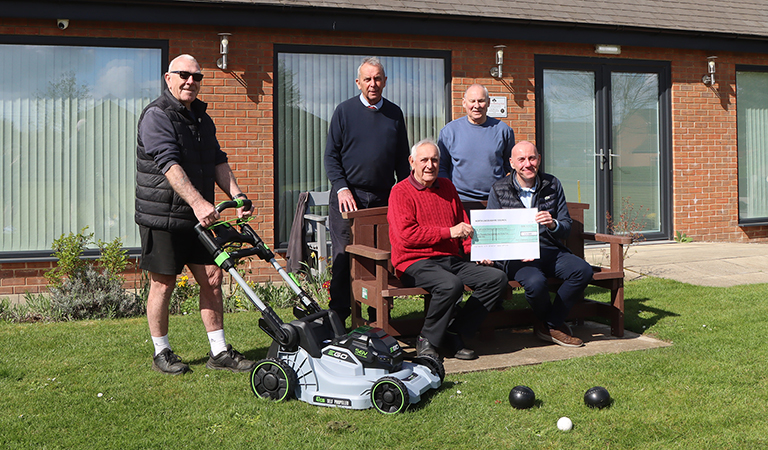 Five men, two sat on a bench holding a big cheque and one with a lawnmower