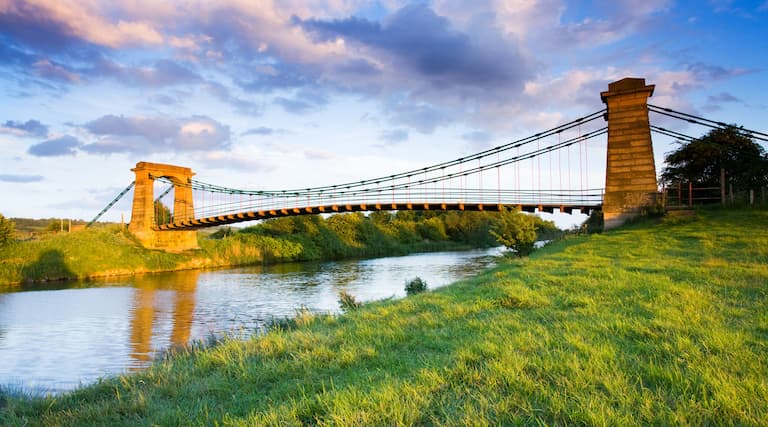 Horkstow suspension bridge seen from the river bank with a blue sky and white clouds beyond