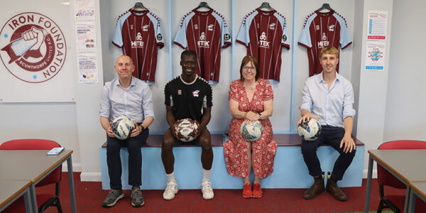 Cllr Rob Waltham, Scunthorpe United player, Cllr Julie Reed and Cllr Josh Walshe sat in the SUFC changing rooms holding a ball each.